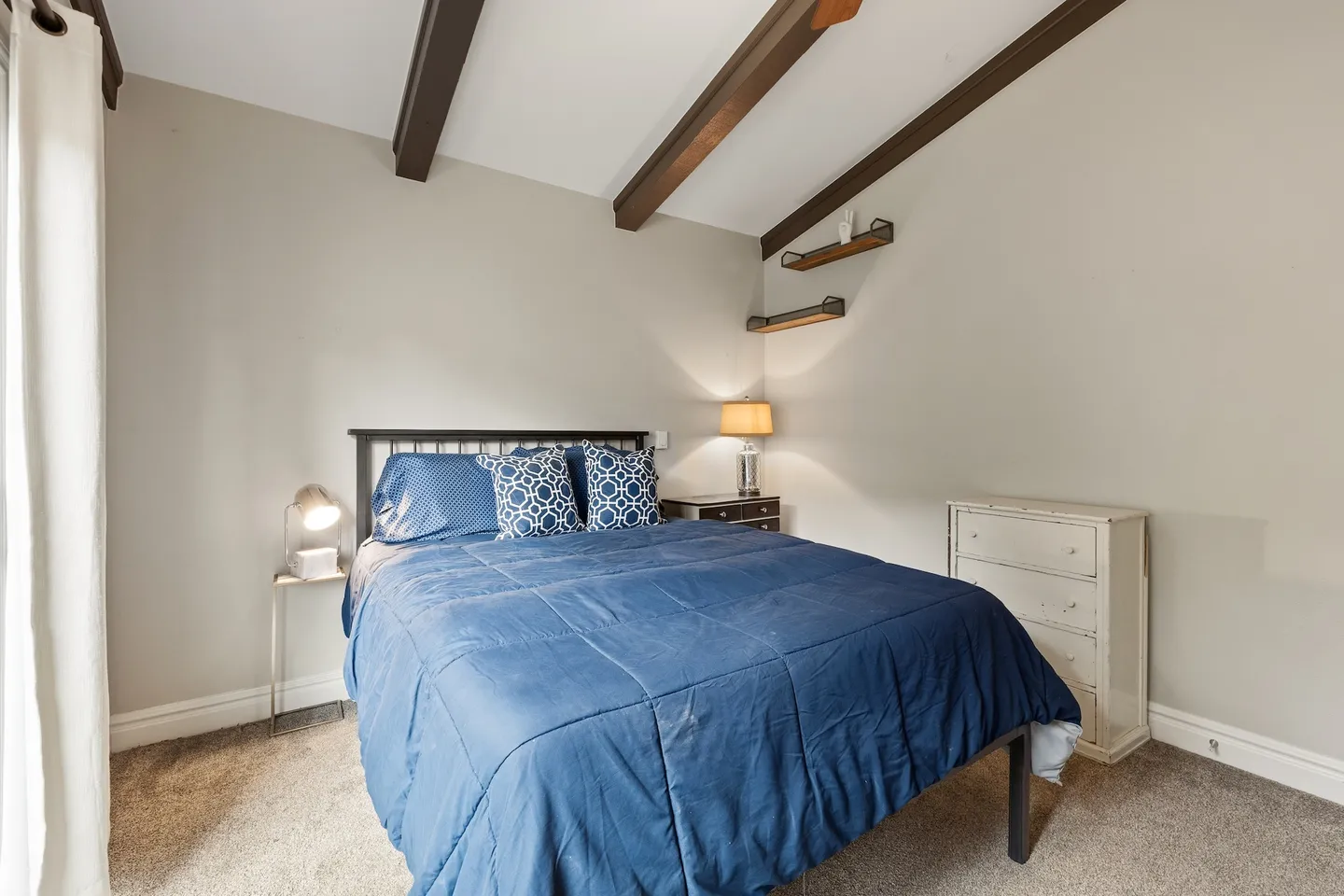 Bedroom with a bed made with blue bedding, vaulted ceiling with dark wood beams, and carpet flooring.