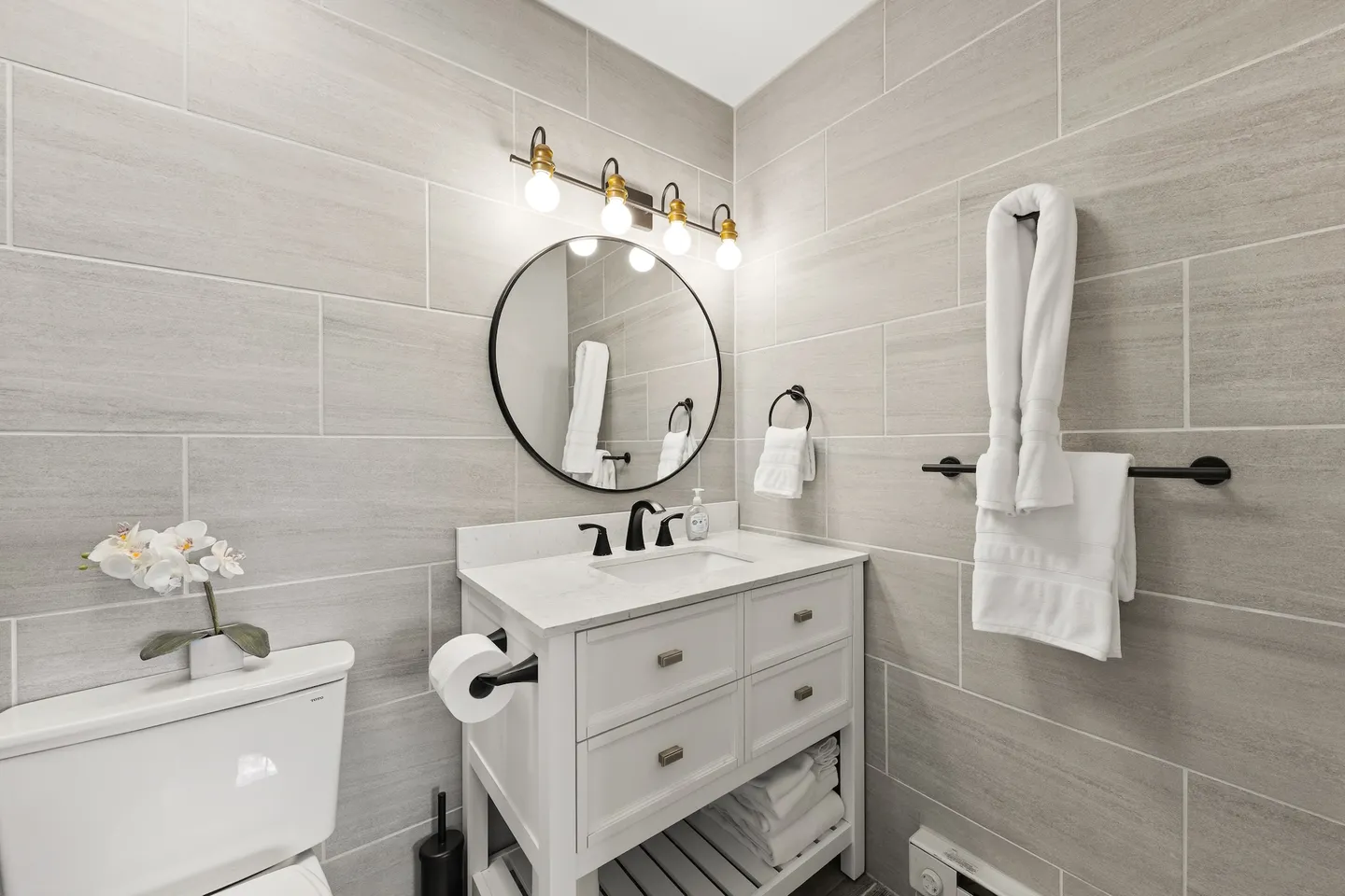 Bathroom with light gray tiled walls, a white vanity with black hardware, a round mirror, and a toilet.