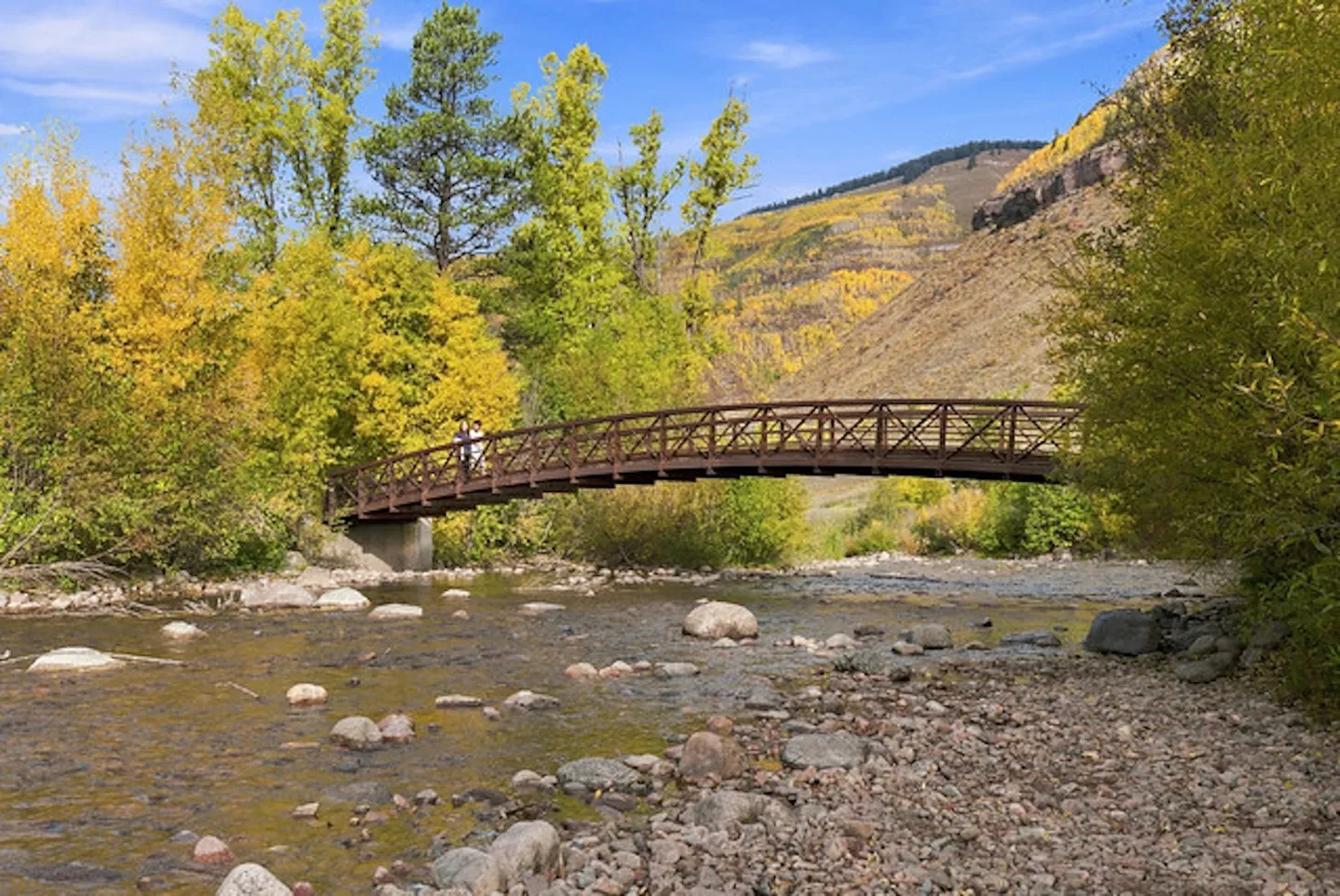 Arched pedestrian bridge crossing over a rocky, shallow river surrounded by autumn trees and mountains.