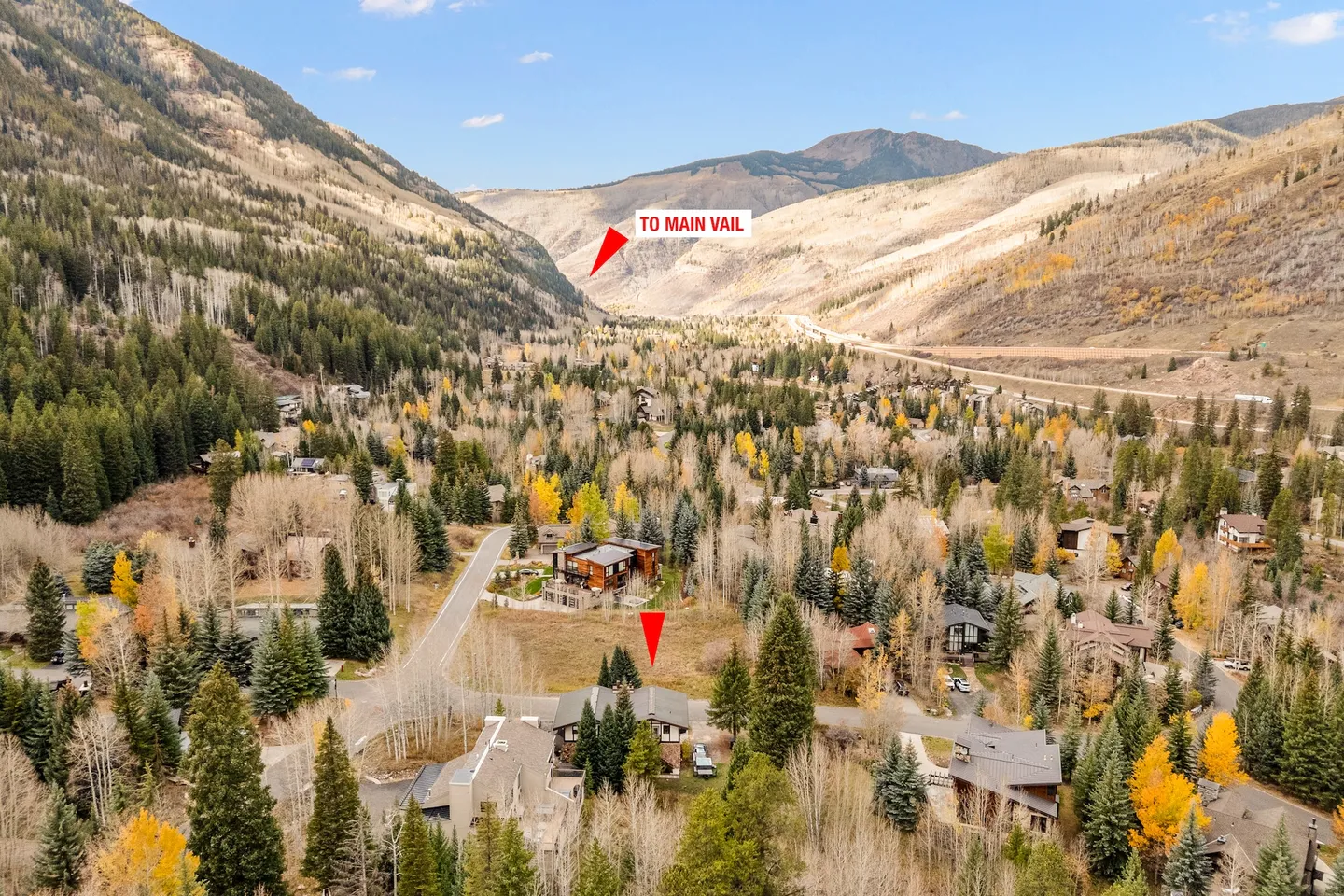 Aerial view of a mountain valley neighborhood surrounded by evergreen and autumn trees, with red directional arrows and text overlay indicating the home location and direction to Main Vail.