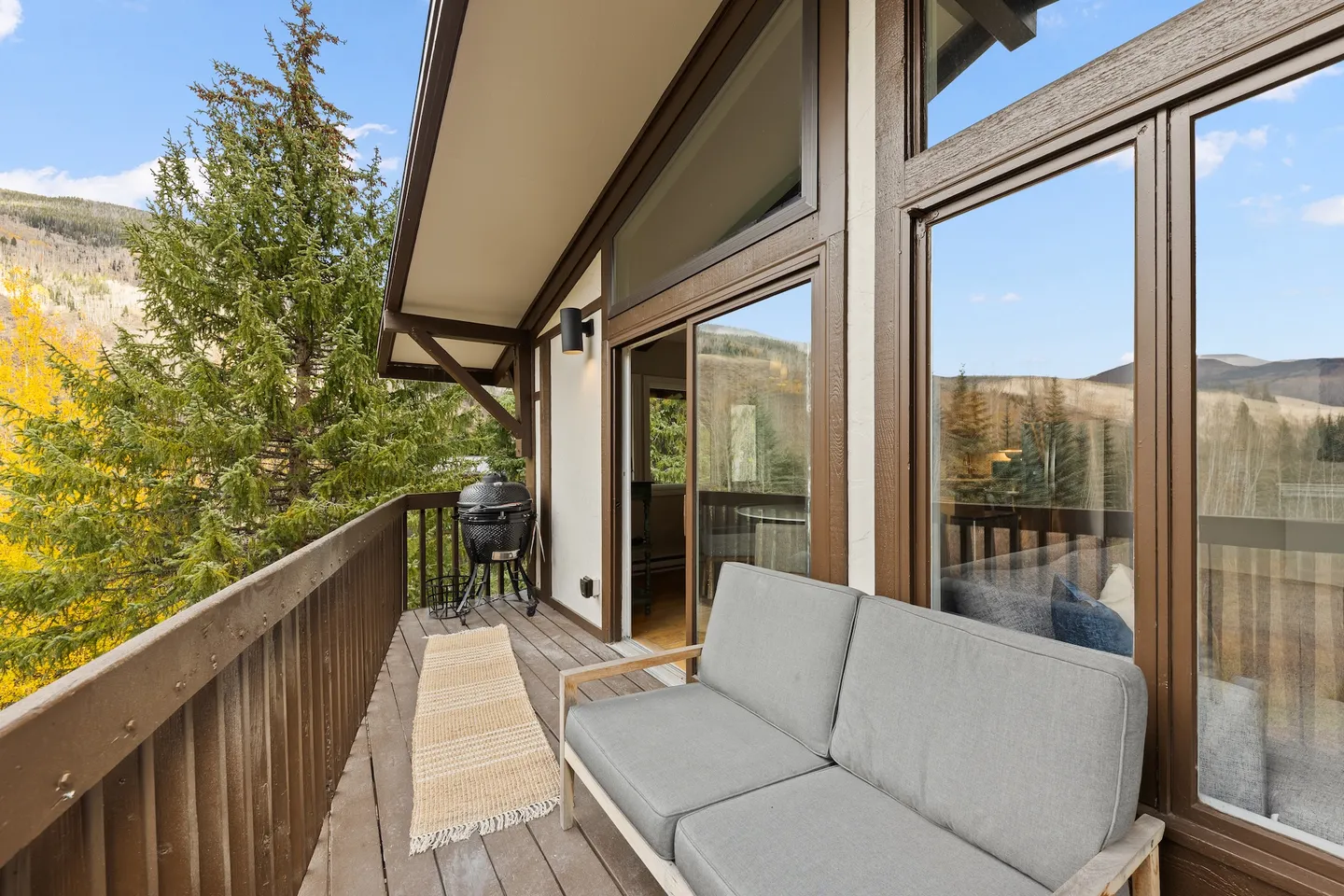 Balcony with a gray cushioned love seat, a runner rug, and a ceramic charcoal grill overlooking trees and mountains.