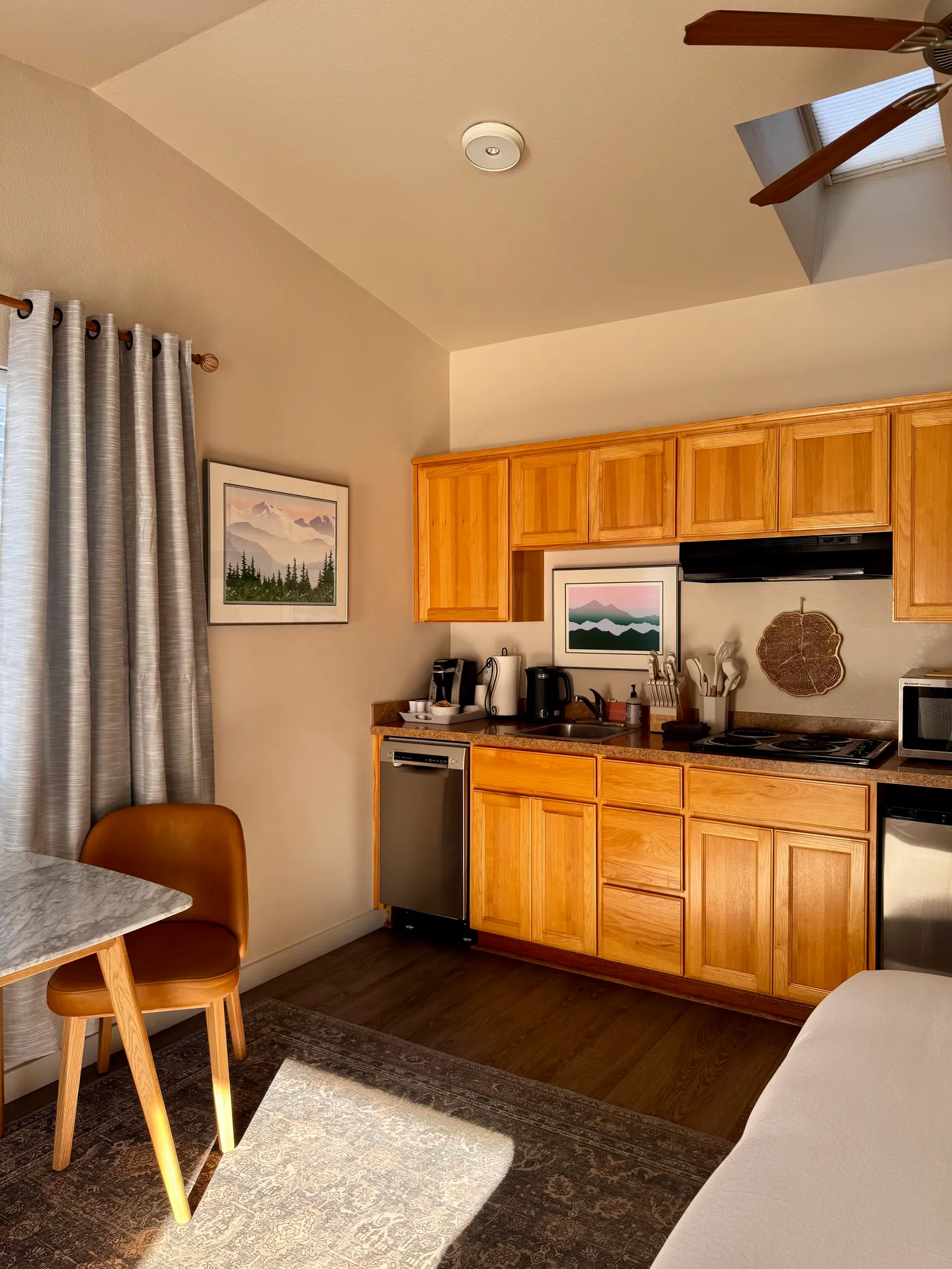 Kitchenette with light wood cabinets, countertop appliances, a small dining table with a chair, and framed art on the walls.
