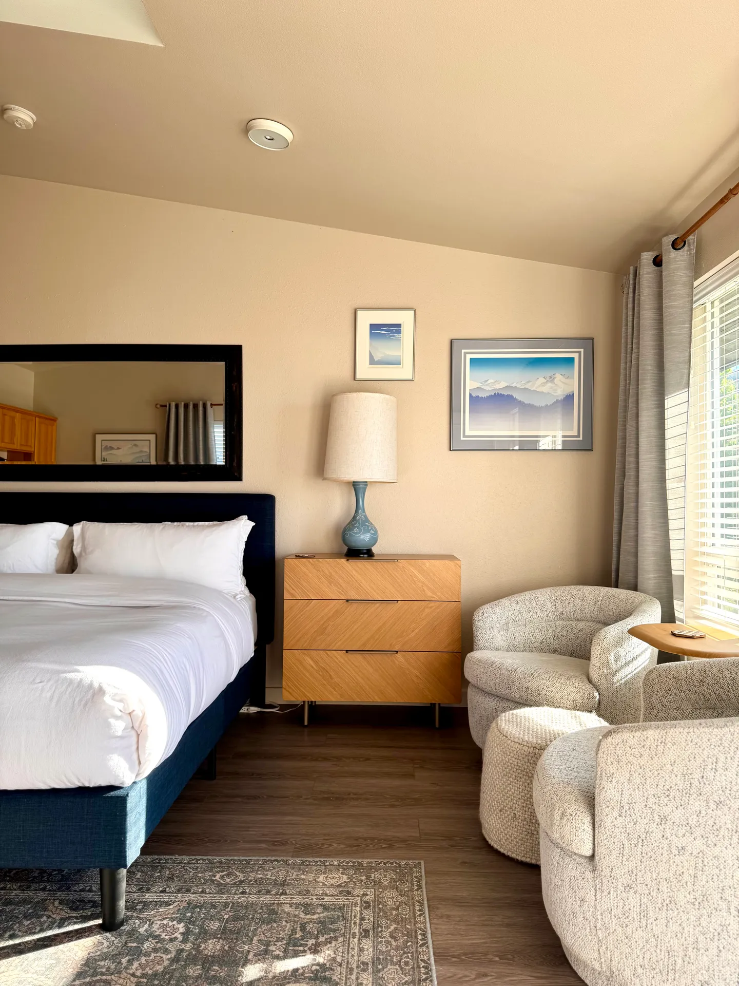 Bedroom featuring a bed with white linens, a nightstand with a lamp, a mirror, and two upholstered swivel armchairs near a window.