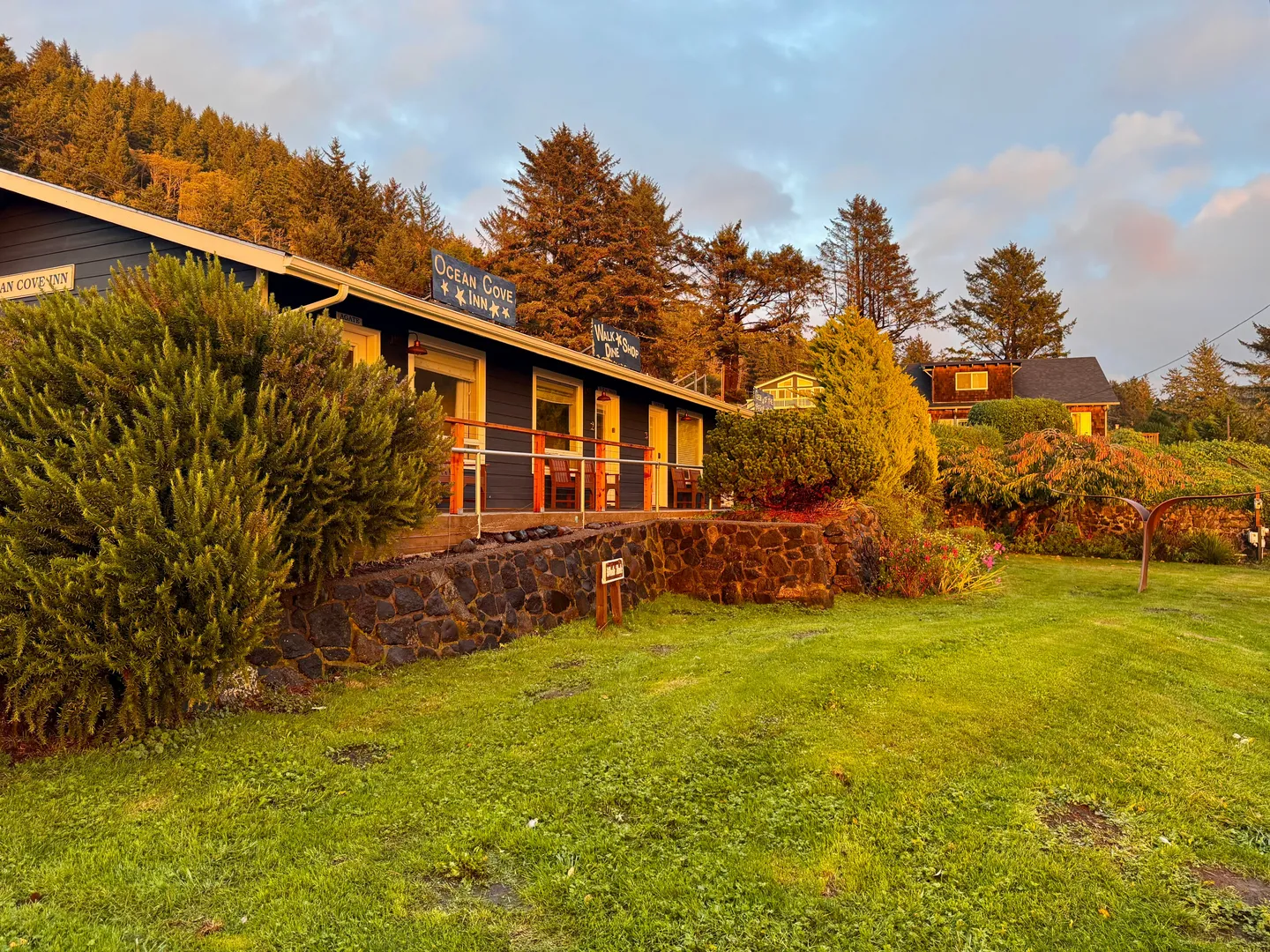 Exterior of Ocean Cove Inn with blue siding, stone retaining wall, and front lawn surrounded by forested hillside.