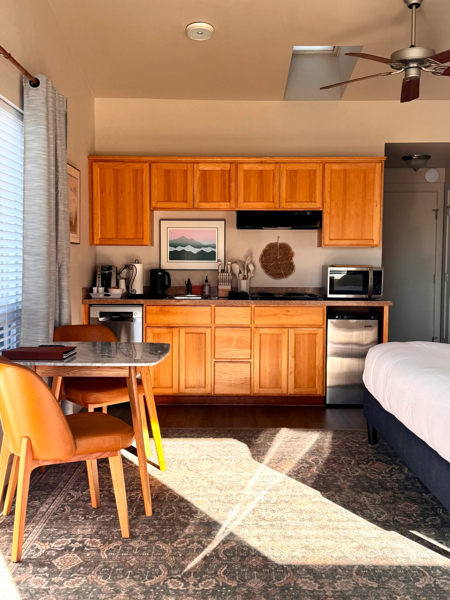Studio interior showing a small kitchen with wooden cabinets, a compact dining table with chairs, and the edge of a bed over a patterned rug.
