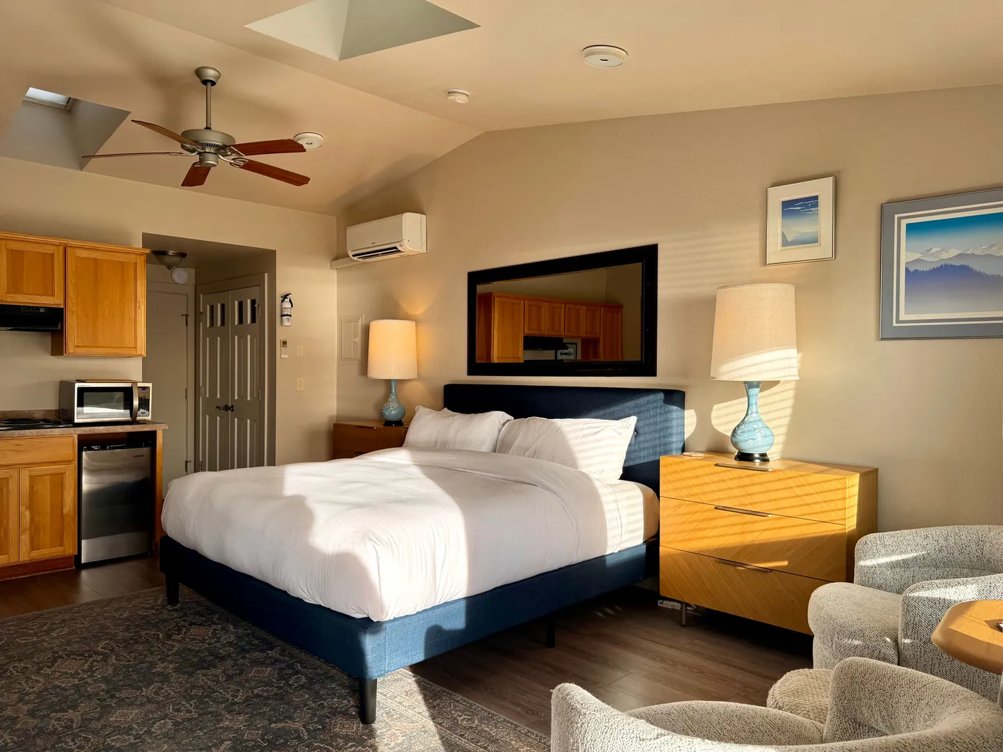 Studio bedroom suite with a blue upholstered bed, skylight ceiling, bedside tables, and a visible kitchenette area.