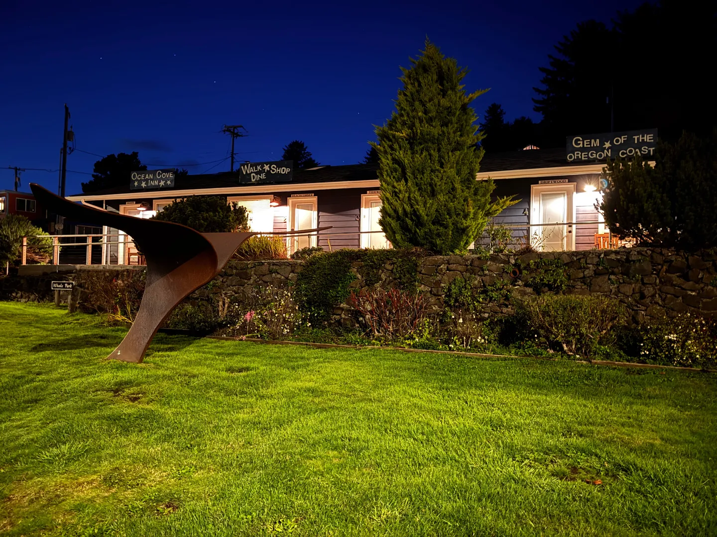 Night exterior view of a single-story inn behind a green lawn, rock retaining wall, and a large metal whale tail sculpture.