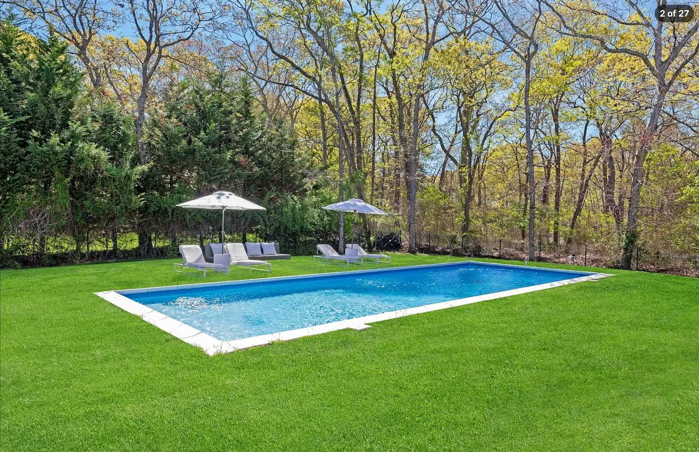A rectangular swimming pool surrounded by a large green lawn, lounge chairs, patio umbrellas, and a dense tree line under a blue sky.