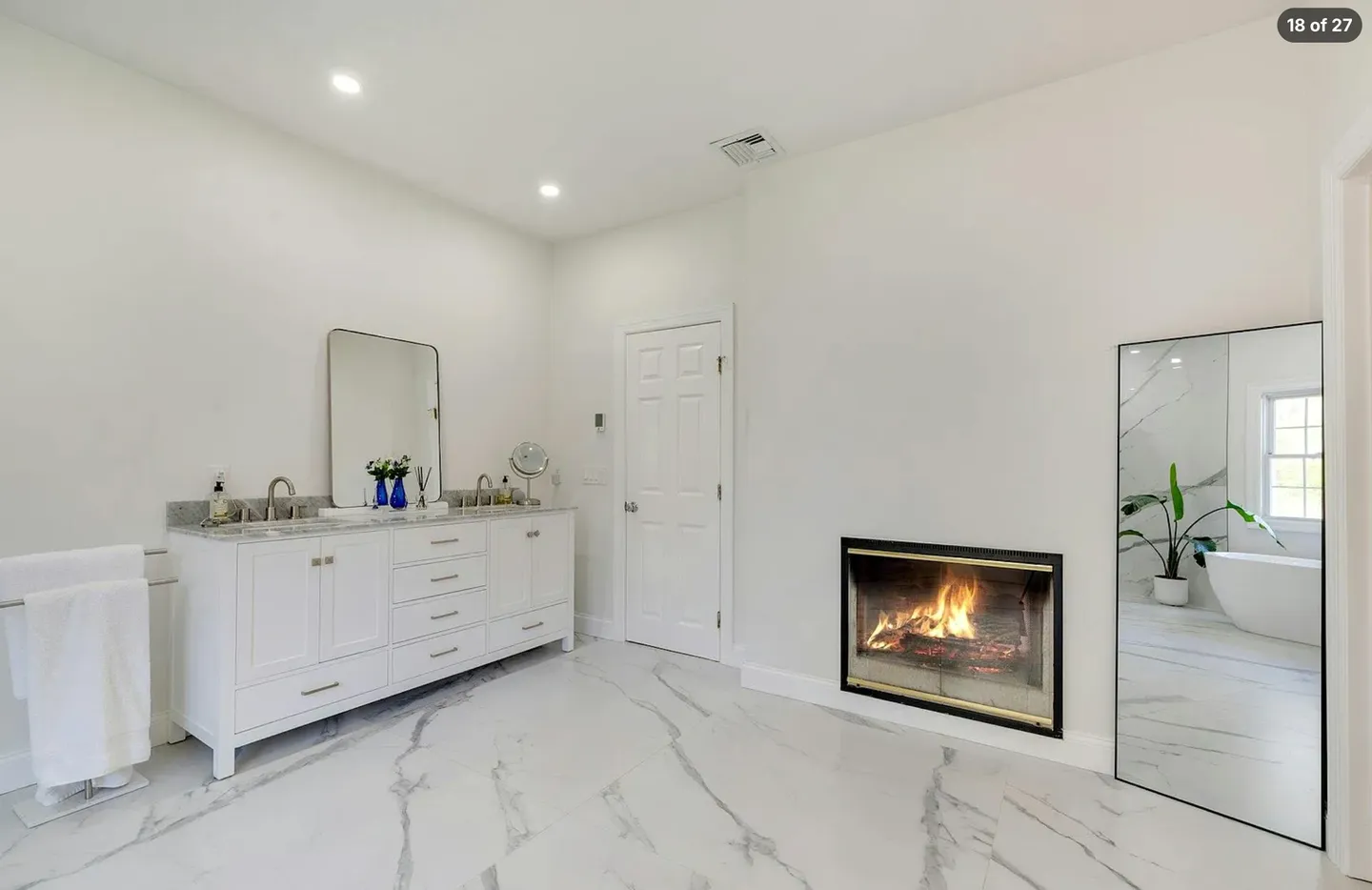 Bathroom with marble-look tile floors, a white vanity with sinks, a lit modern fireplace, and a full-length mirror reflecting a bathtub.