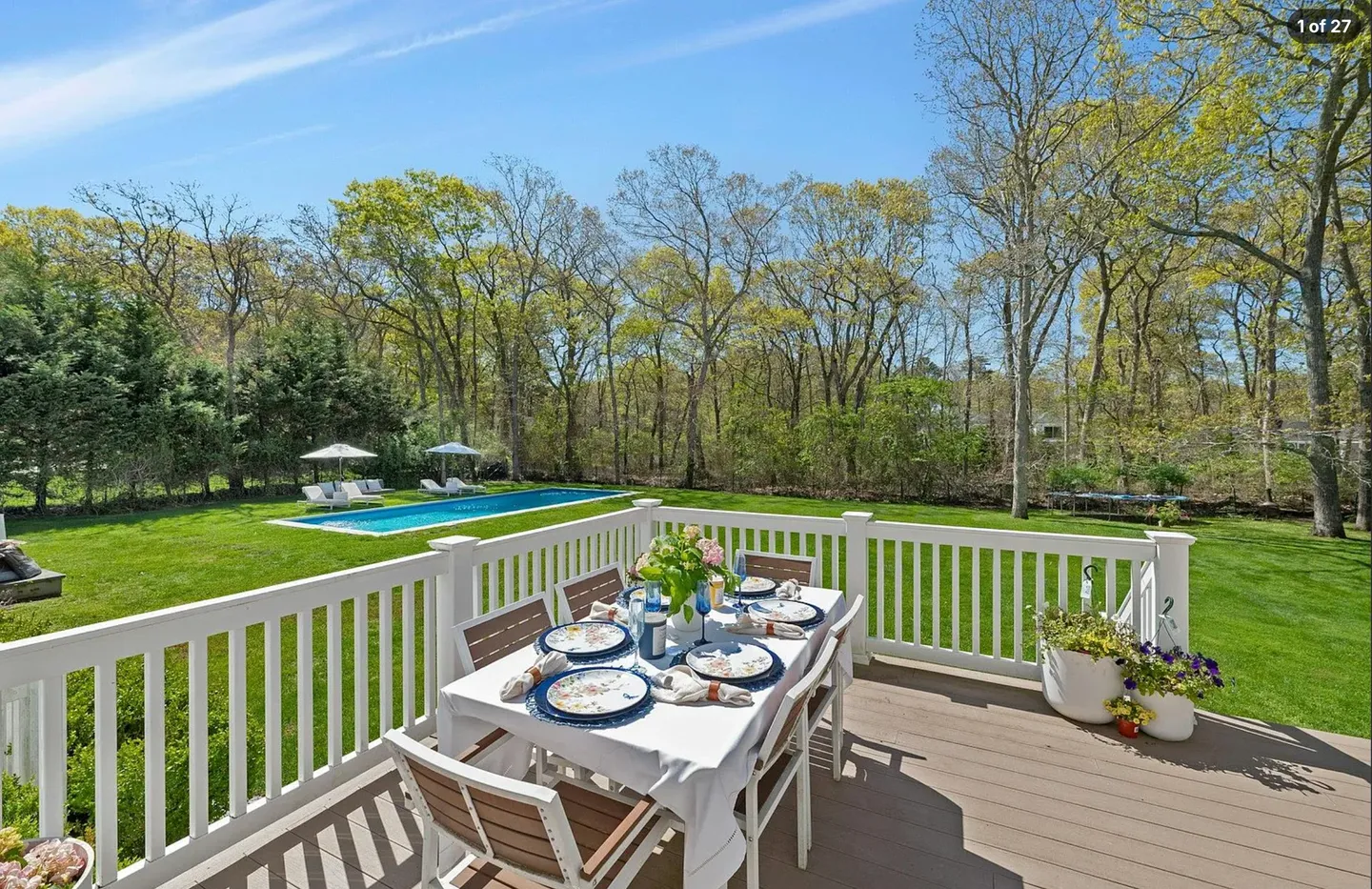 Elevated wooden deck with a set dining table and chairs overlooking a green lawn, swimming pool, and wooded area under a blue sky.