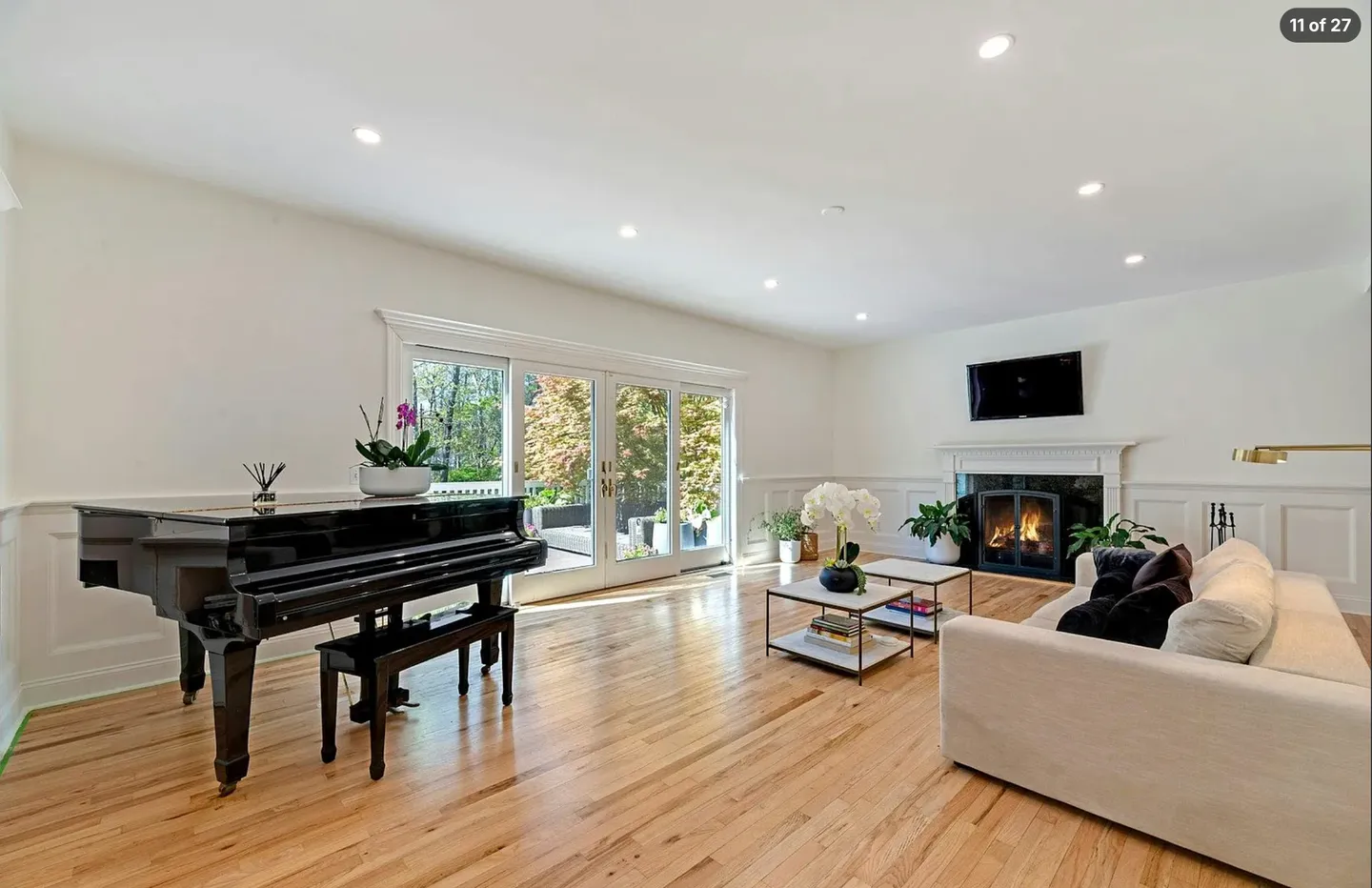 Bright living room featuring light hardwood floors, a black grand piano, a fireplace, and glass doors opening to an outdoor patio.