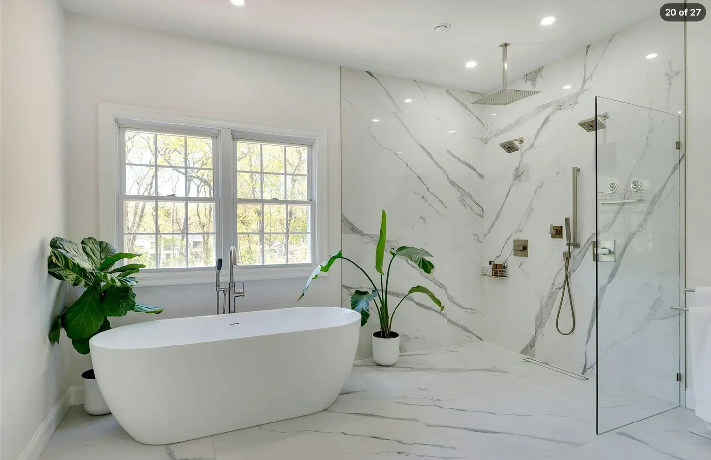Bright modern bathroom featuring a freestanding oval soaking tub beside windows and an open walk-in shower with marble-style tile walls.