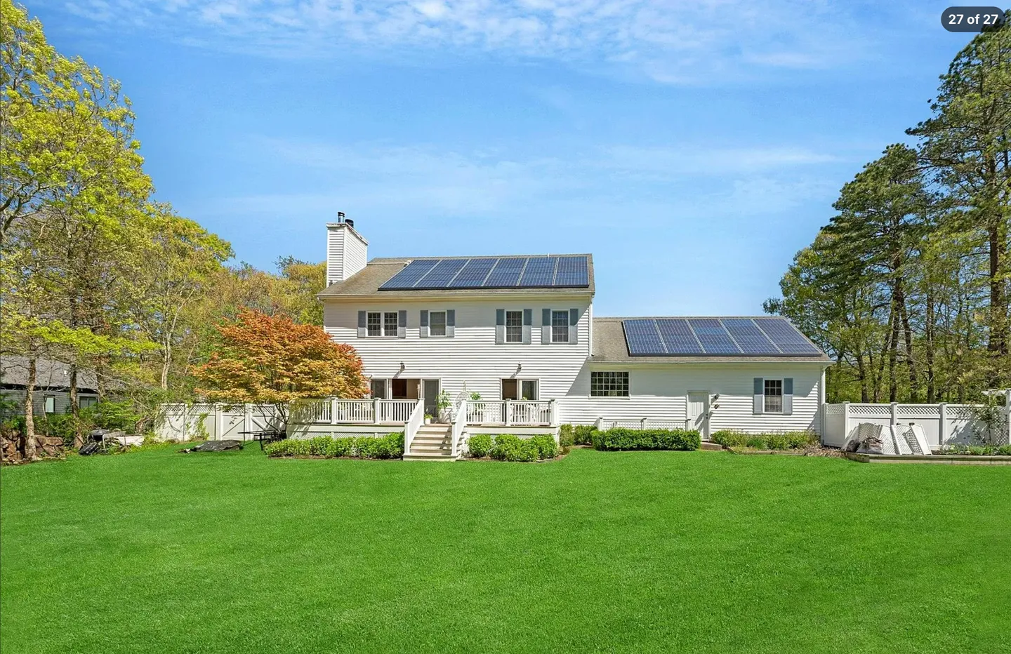 Rear exterior view of a two-story white house with solar panels on the roof and a spacious green lawn.