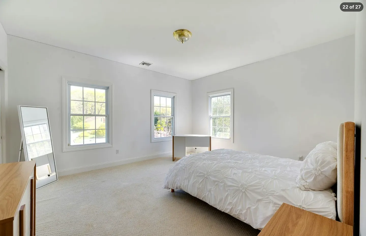 Bedroom with carpeted floors, white walls, three windows, a bed with white textured bedding, a full-length mirror, and a small desk in the corner.