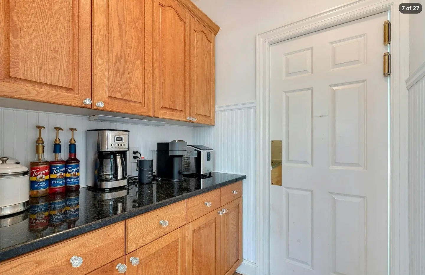 Dedicated coffee station with light wood cabinetry, dark countertop, multiple coffee makers, syrup bottles, and a white door adjacent to white beadboard walls.