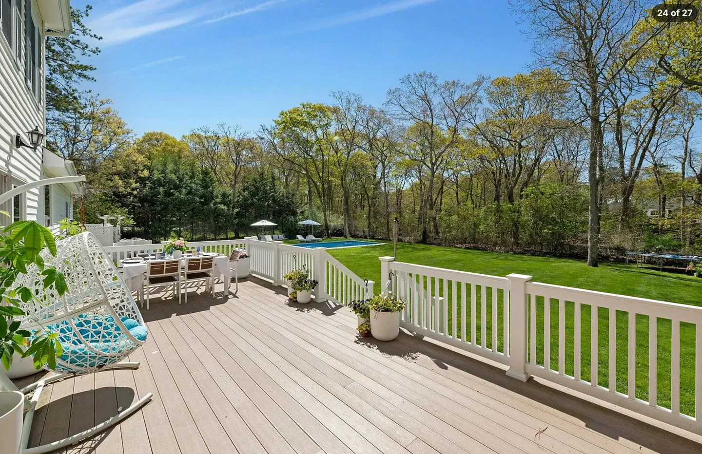 Elevated wooden deck with outdoor dining table, hanging chair, and white railings overlooking a large lawn and swimming pool surrounded by trees.