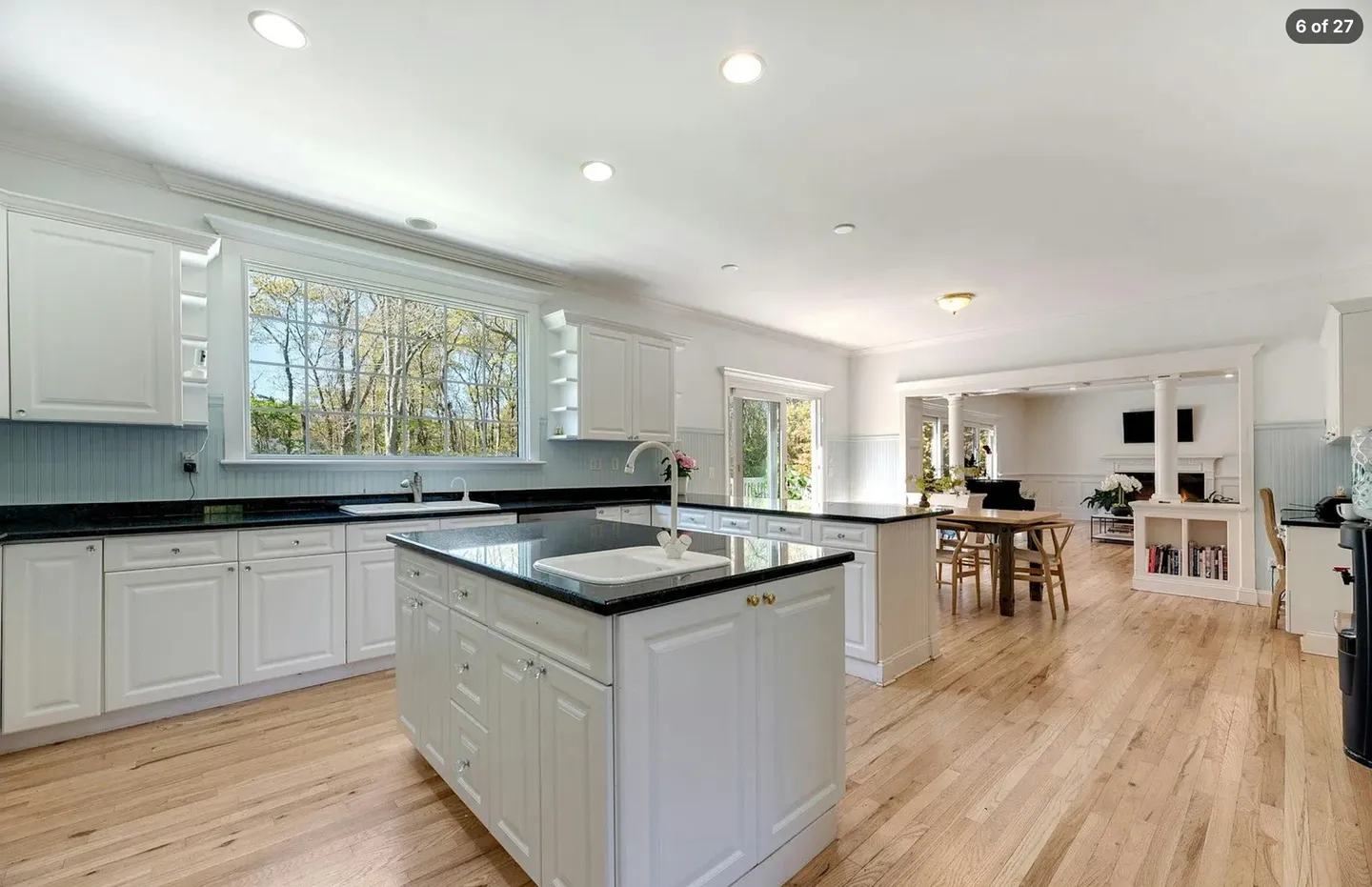 Spacious kitchen with white cabinetry, dark countertops, a center island with a prep sink, and light wood floors opening to a dining and living area.