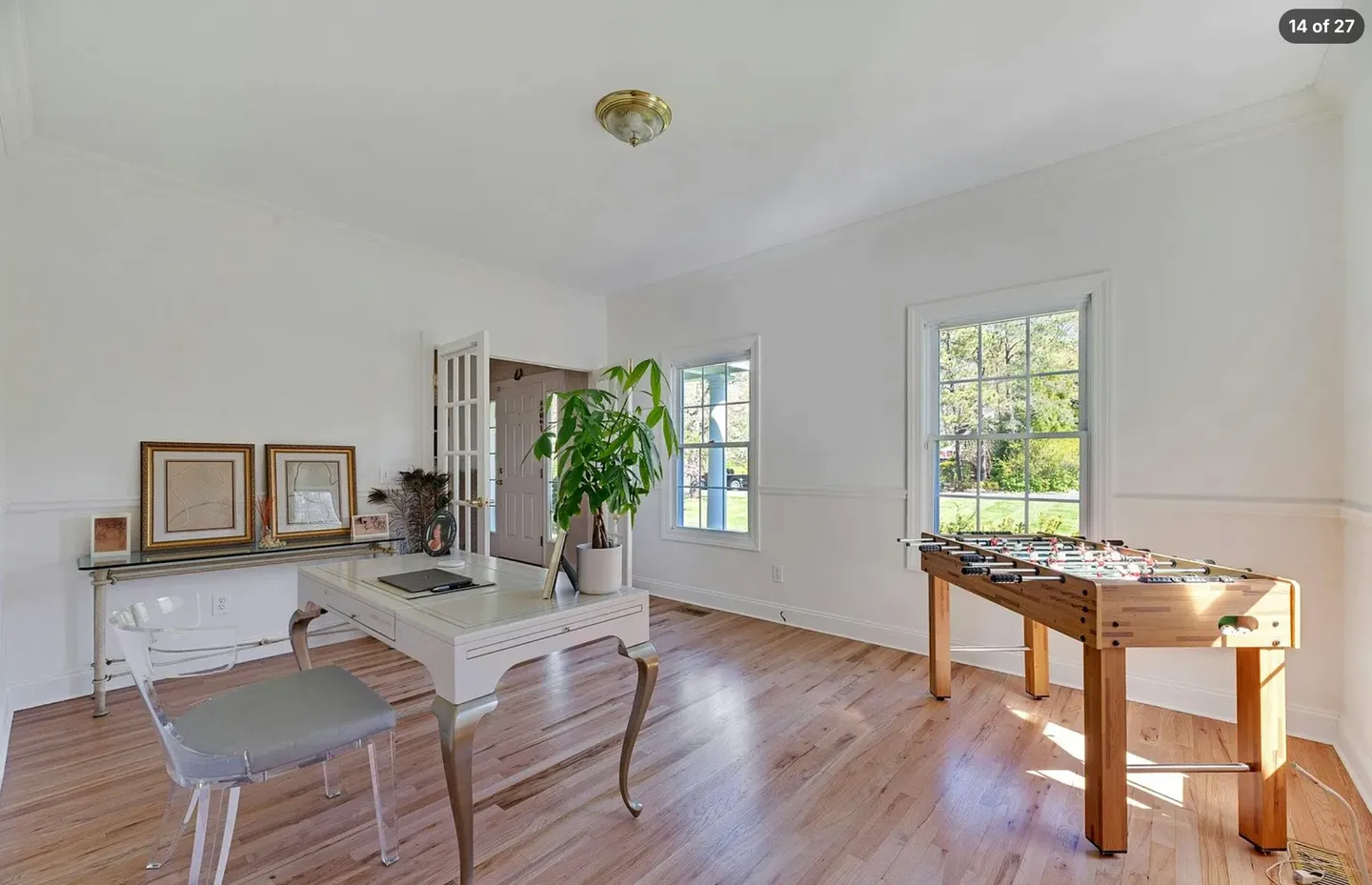 Bright room with wood-style flooring featuring a white desk with a clear chair, a console table with framed pictures, and a wooden foosball table by the window.