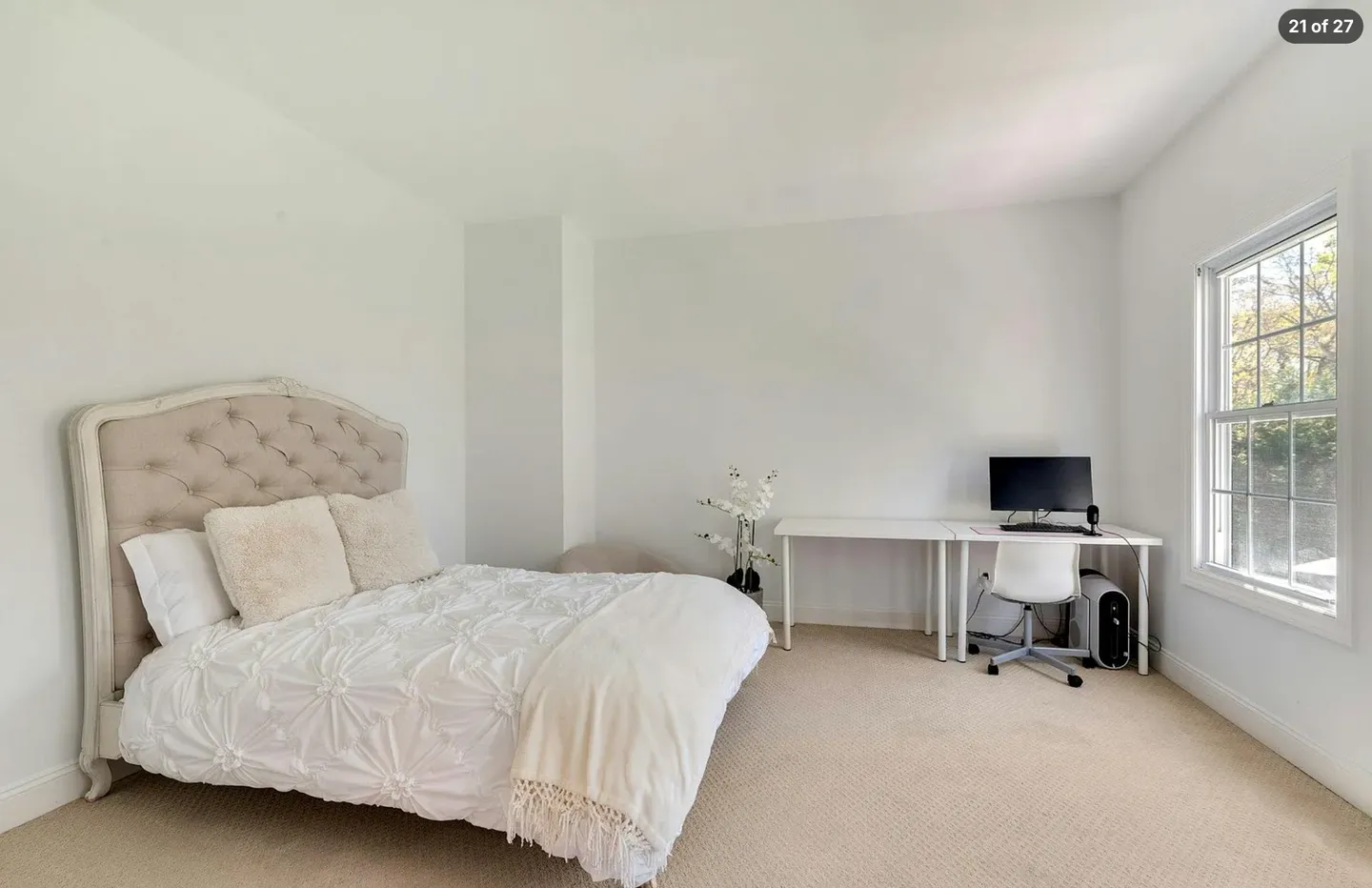 Bright bedroom with carpet, a tufted headboard on a made bed, and a desk workspace next to a window.