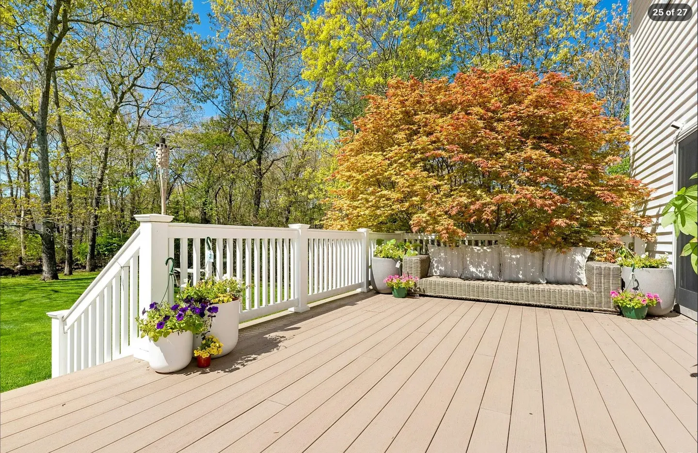Outdoor raised wooden deck with white railings, a wicker sofa, potted flowers, and a large colorful tree under a clear blue sky.