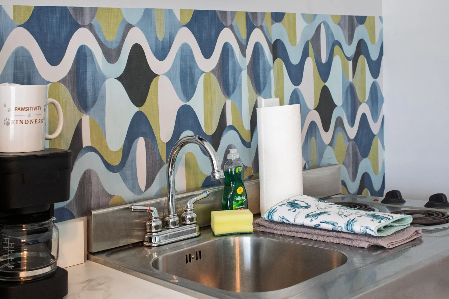 Close-up of a kitchen sink area with stainless steel counter, coffee maker, dish soap, sponge, paper towels, and a retro wavy blue and green backsplash.