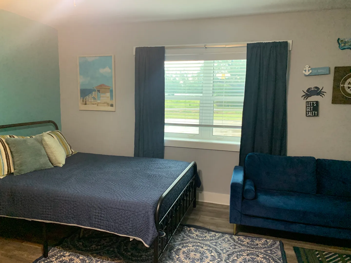 Bedroom featuring a metal bed frame with navy bedding, a blue sofa, and a window with navy curtains.