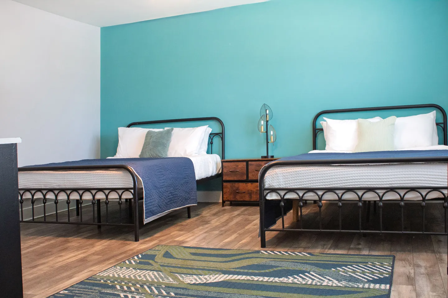 Bedroom featuring two black metal frame beds against a bright teal accent wall with a wooden nightstand in between.