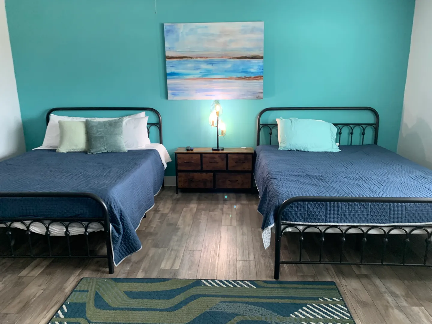 Bedroom featuring two beds with black metal frames, blue coverlets, a small dresser between them, and a bright turquoise accent wall.