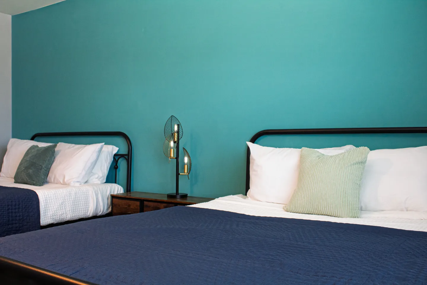 Bedroom featuring two beds with black metal frames and blue bedspreads against a teal accent wall.