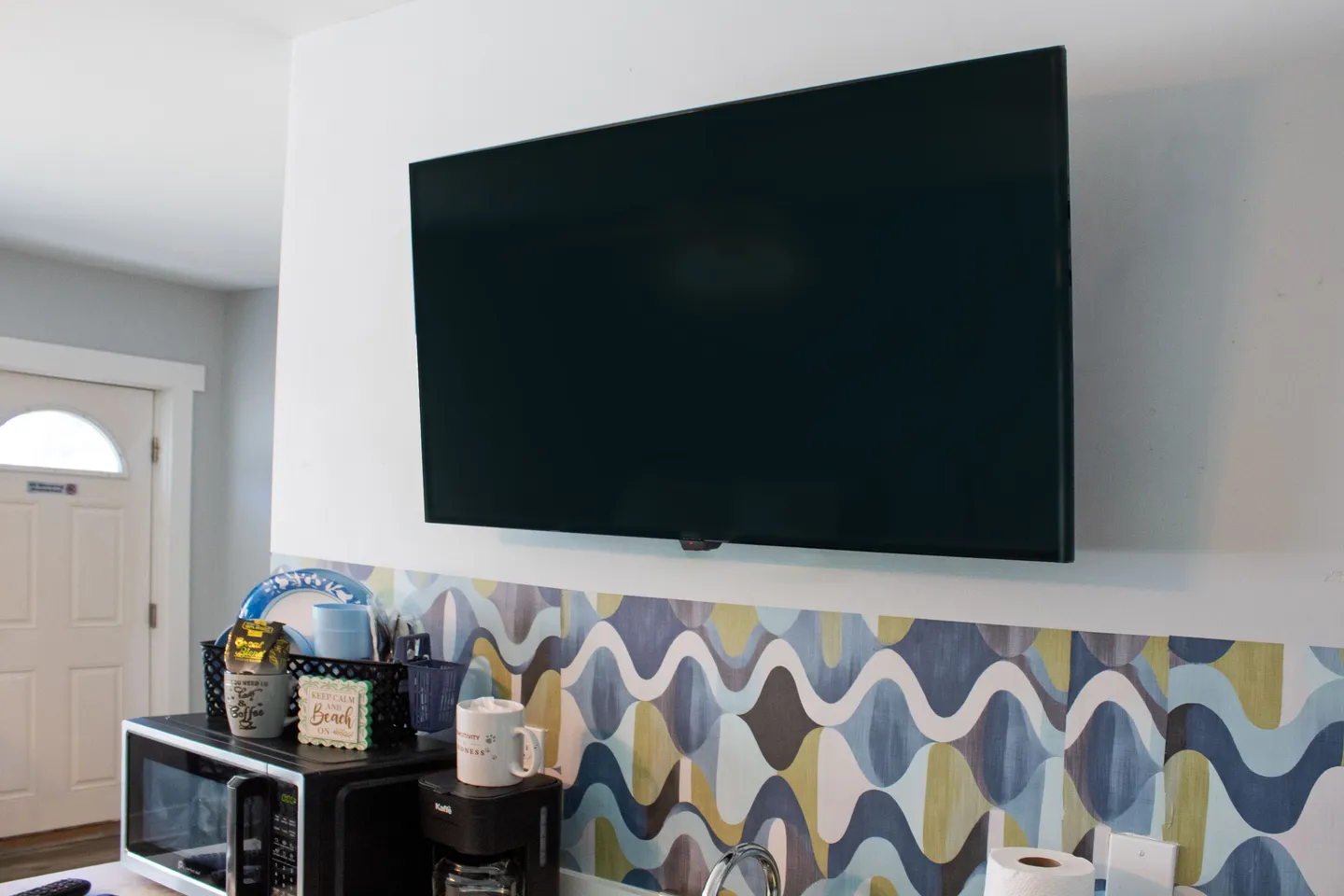 A flat-screen TV mounted on a white wall above a countertop with patterned tile backsplash, a microwave, and coffee maker.