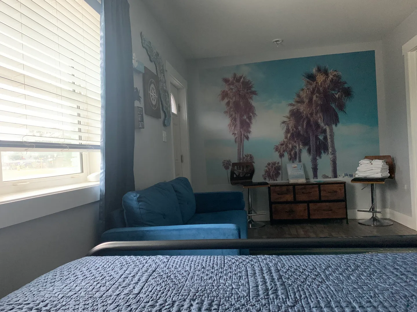 Bedroom and sitting area with a blue couch, window with blinds, palm tree wall mural, and a small counter with barstools.