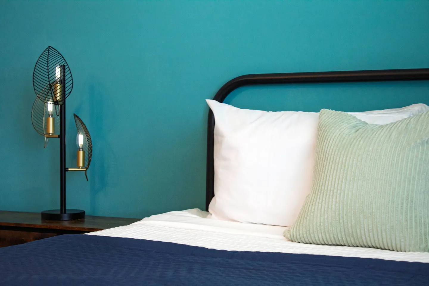 Close-up of a bed with a black metal frame, pillows, and a modern decorative lamp against a teal wall.