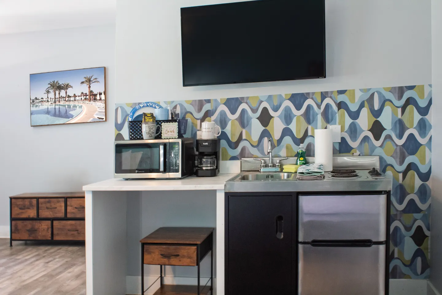 A compact kitchenette setup featuring a sink, mini-fridge, microwave, coffee maker, and wall-mounted TV.