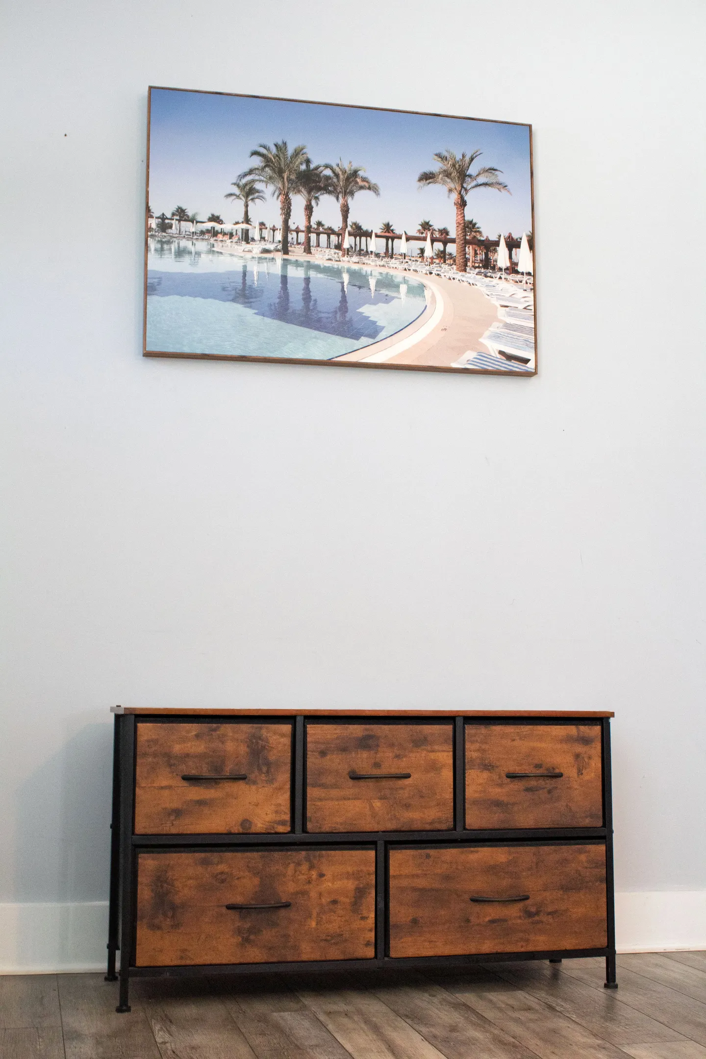 A rustic wood-look and black metal dresser positioned against a plain wall underneath a framed picture of a tropical resort pool.