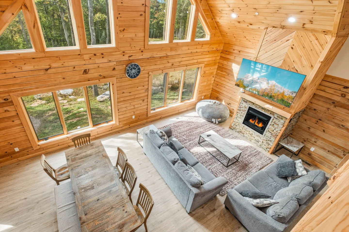 High-angle view of a wood-paneled living room with a fireplace, television, two couches, and a dining table beside large windows.