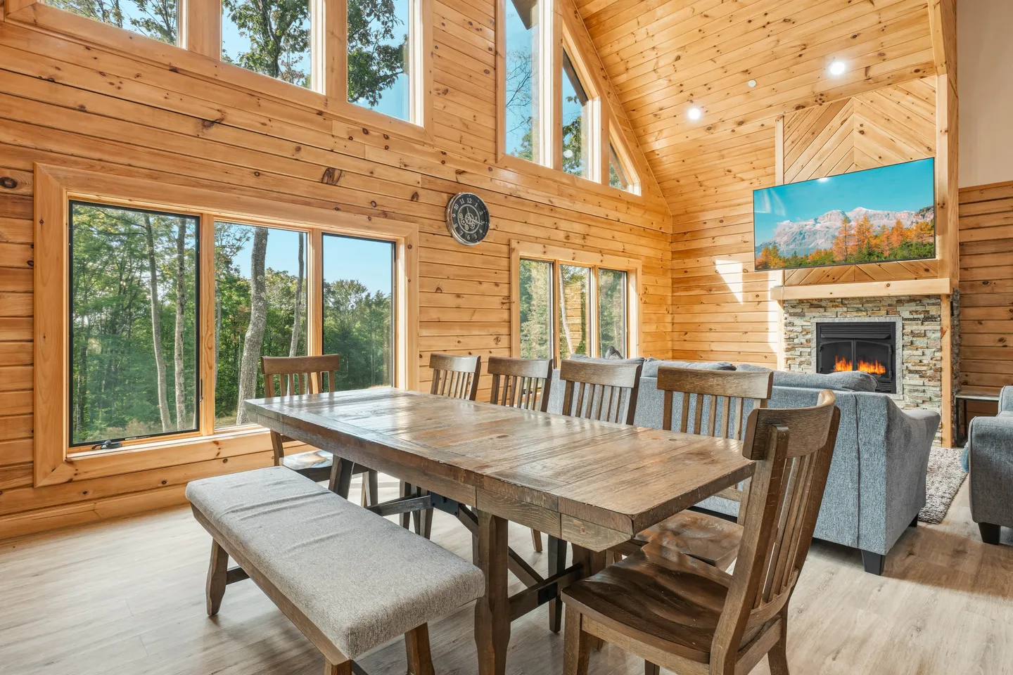 Dining area with a wooden table, bench, and chairs in a log-walled open-plan room with large windows and an adjacent living area with a fireplace.