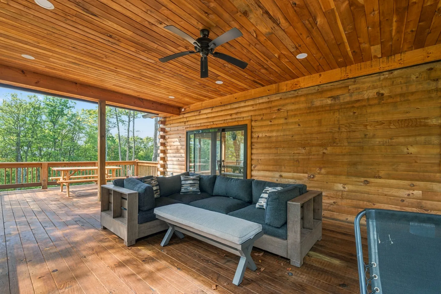 Covered wooden deck featuring a cushioned sectional sofa, bench, ceiling fan, and log siding.