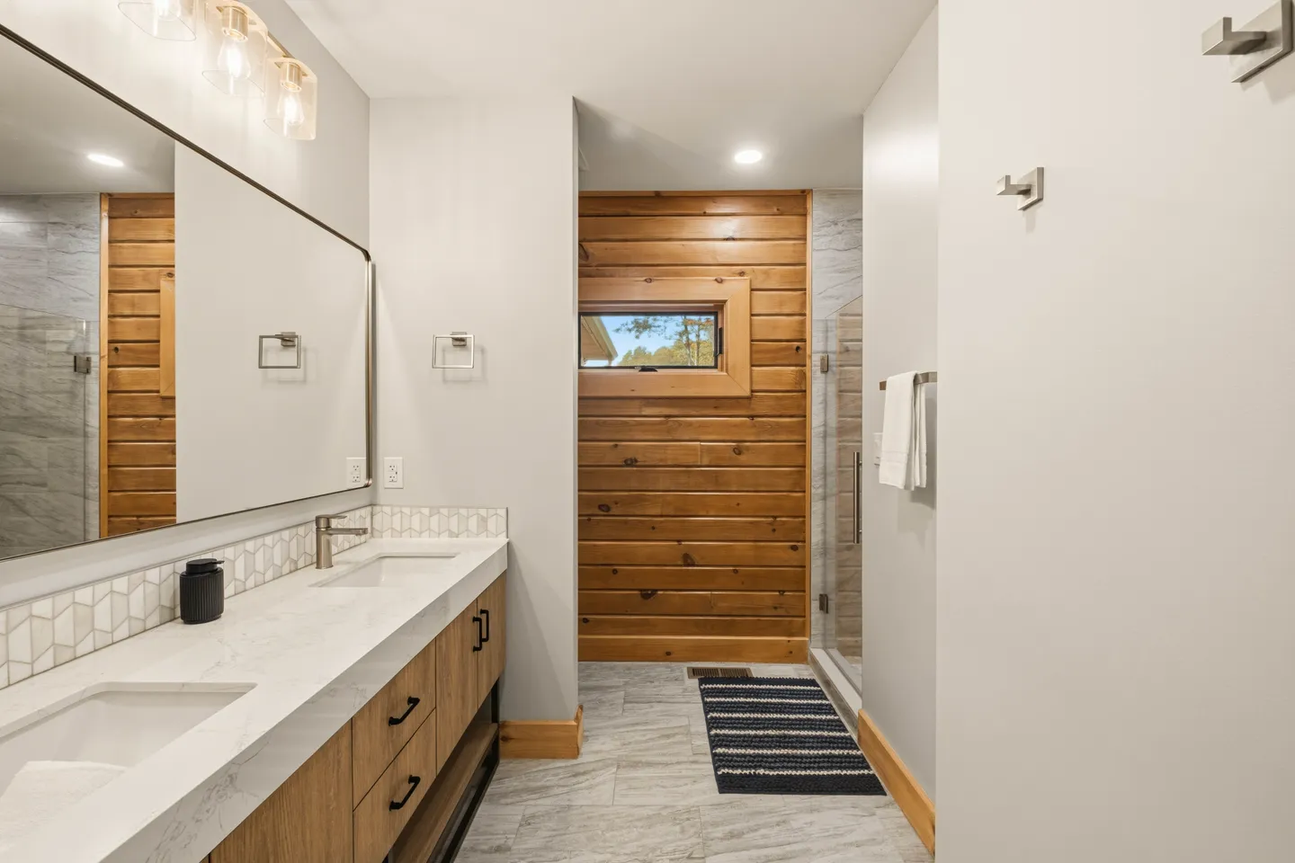 Bathroom with a double-sink vanity, large mirror, tile floor, glass walk-in shower, and a wood-plank accent wall with a small window.