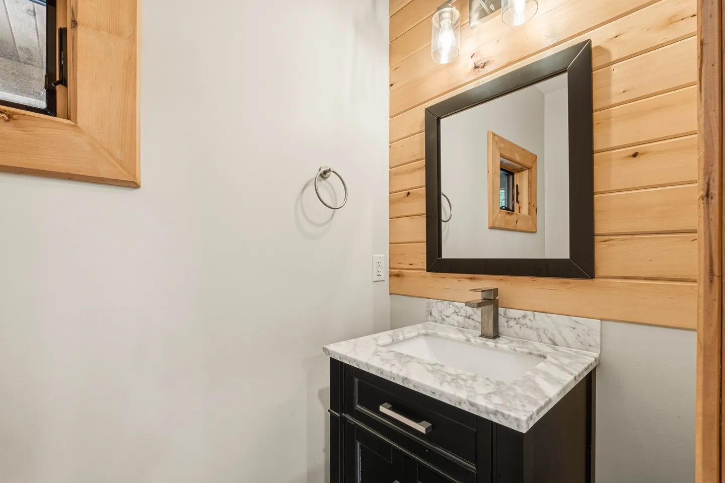 Bathroom vanity with a dark cabinet, marble-look countertop, rectangular framed mirror, and horizontal wood-plank accent wall.