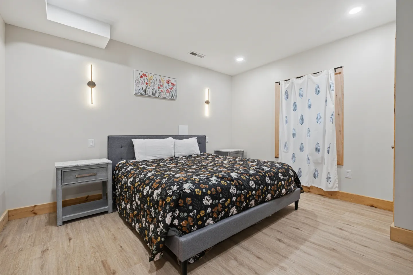 Bedroom with an upholstered bed, floral bedding, two bedside tables, wall sconces, and light wood-look flooring.