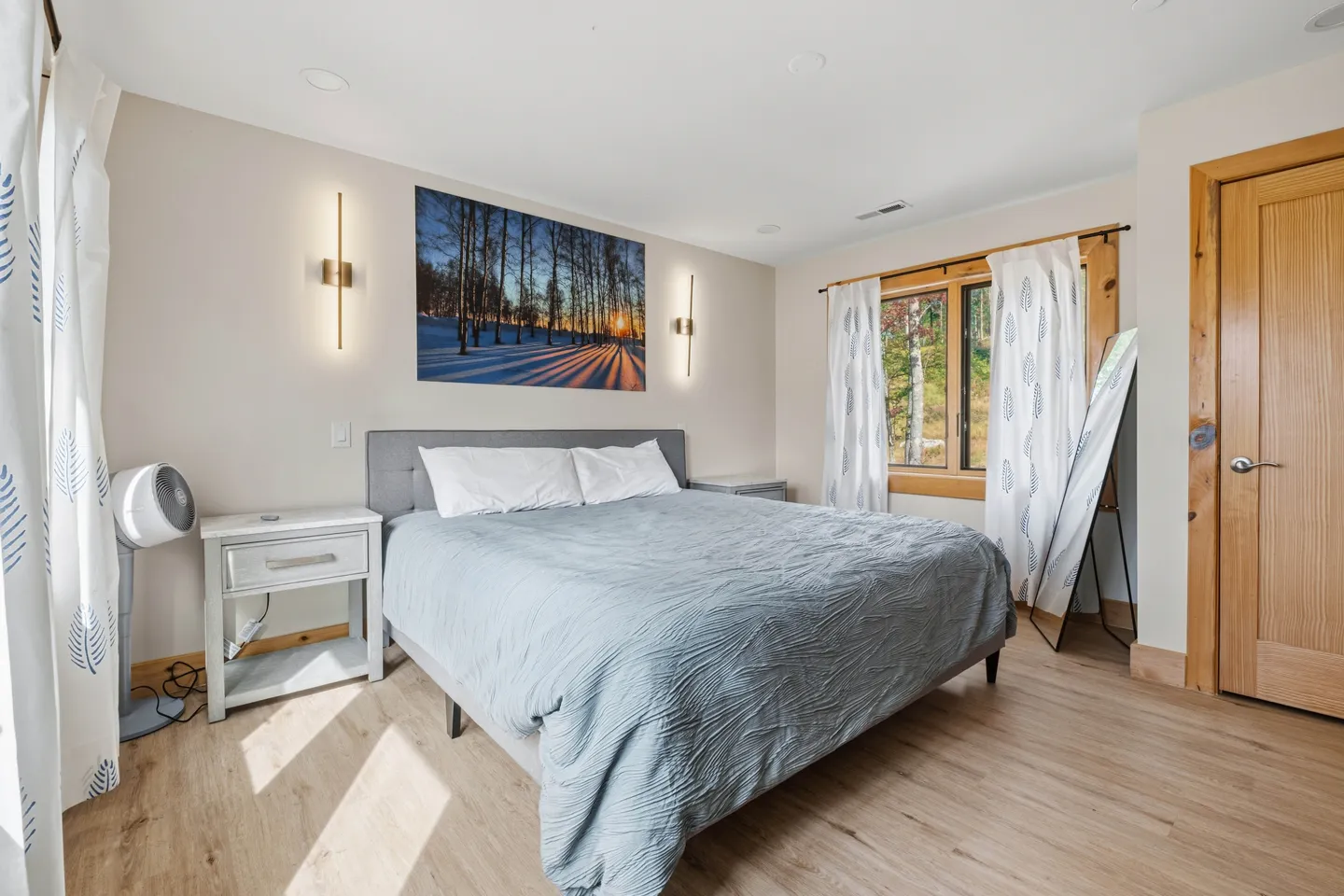 Bedroom with a bed made in blue bedding, modern wall sconces, wood-look flooring, and a window.