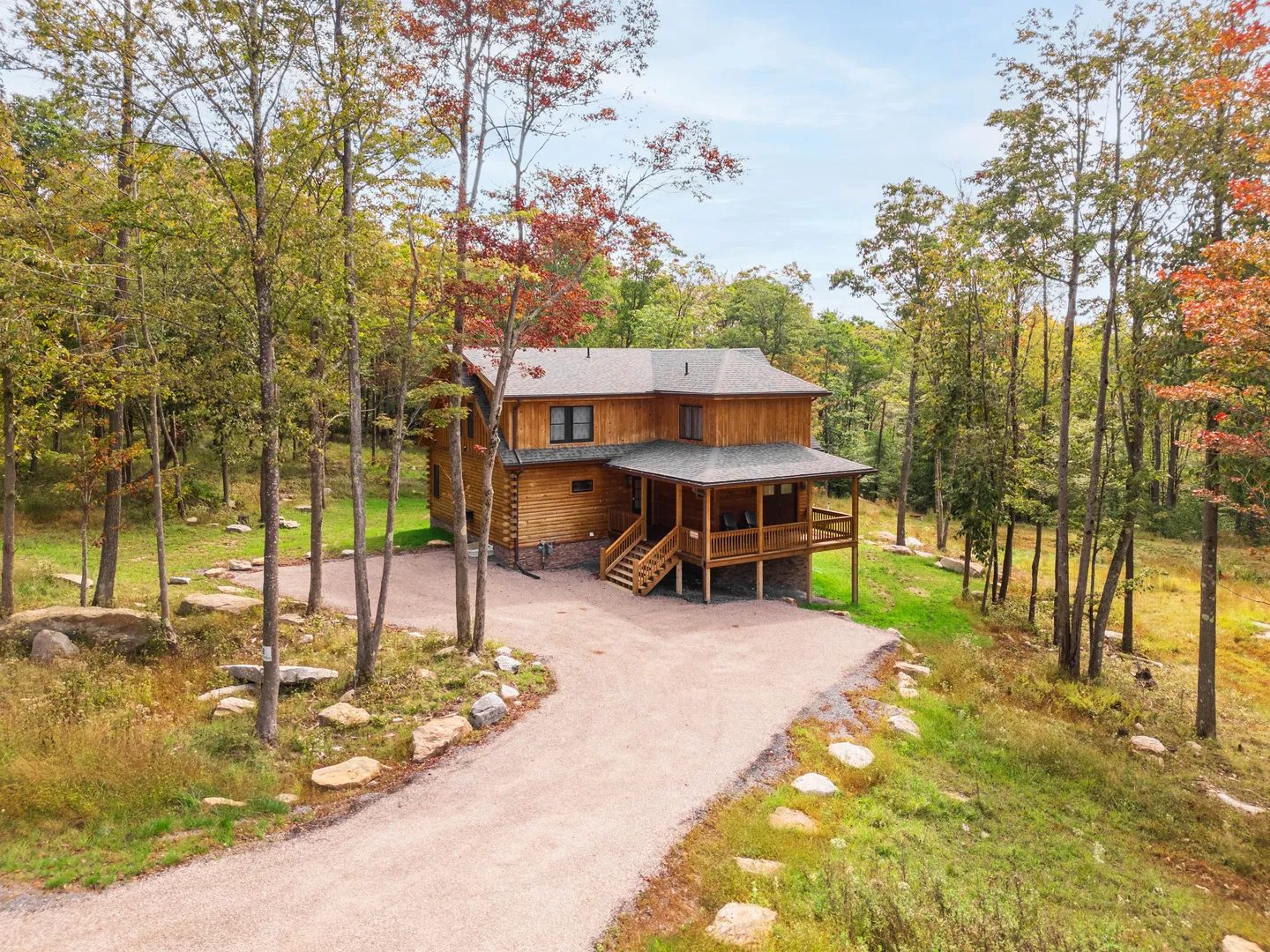 Aerial view of a two-story log home with a covered front porch and gravel driveway surrounded by trees.