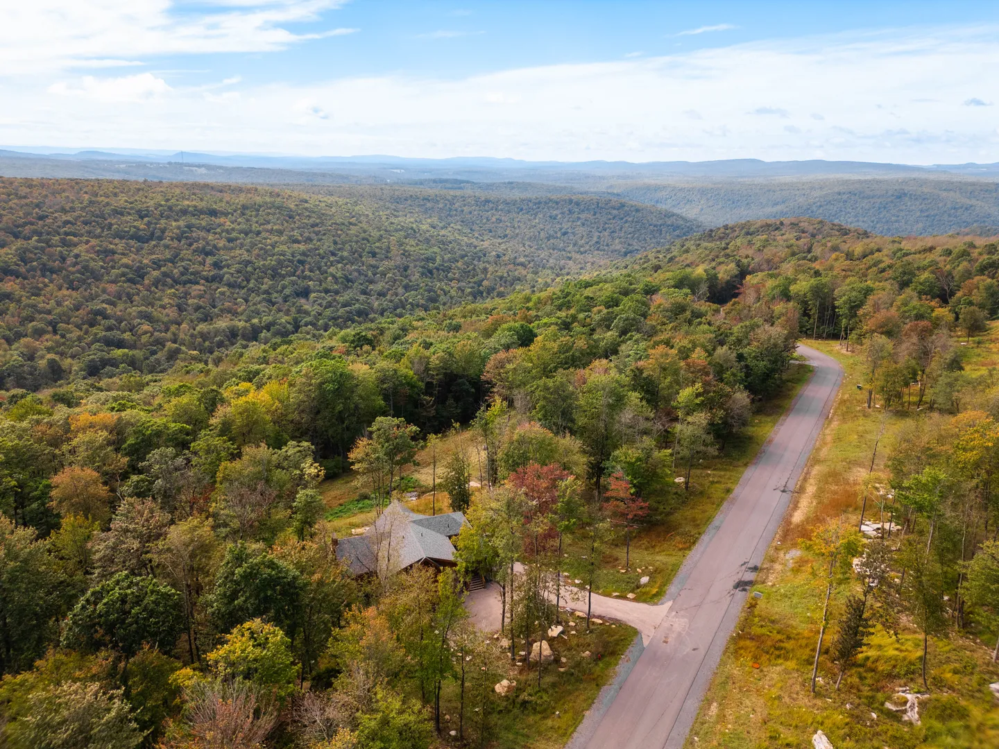 Aerial view of a house nestled in a dense forest alongside a paved road with rolling hills in the background.