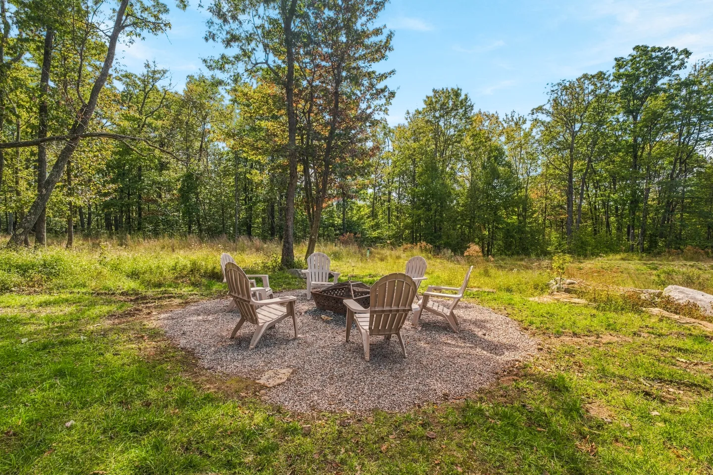 Outdoor fire pit surrounded by Adirondack chairs on a gravel patch bordering a wooded area.