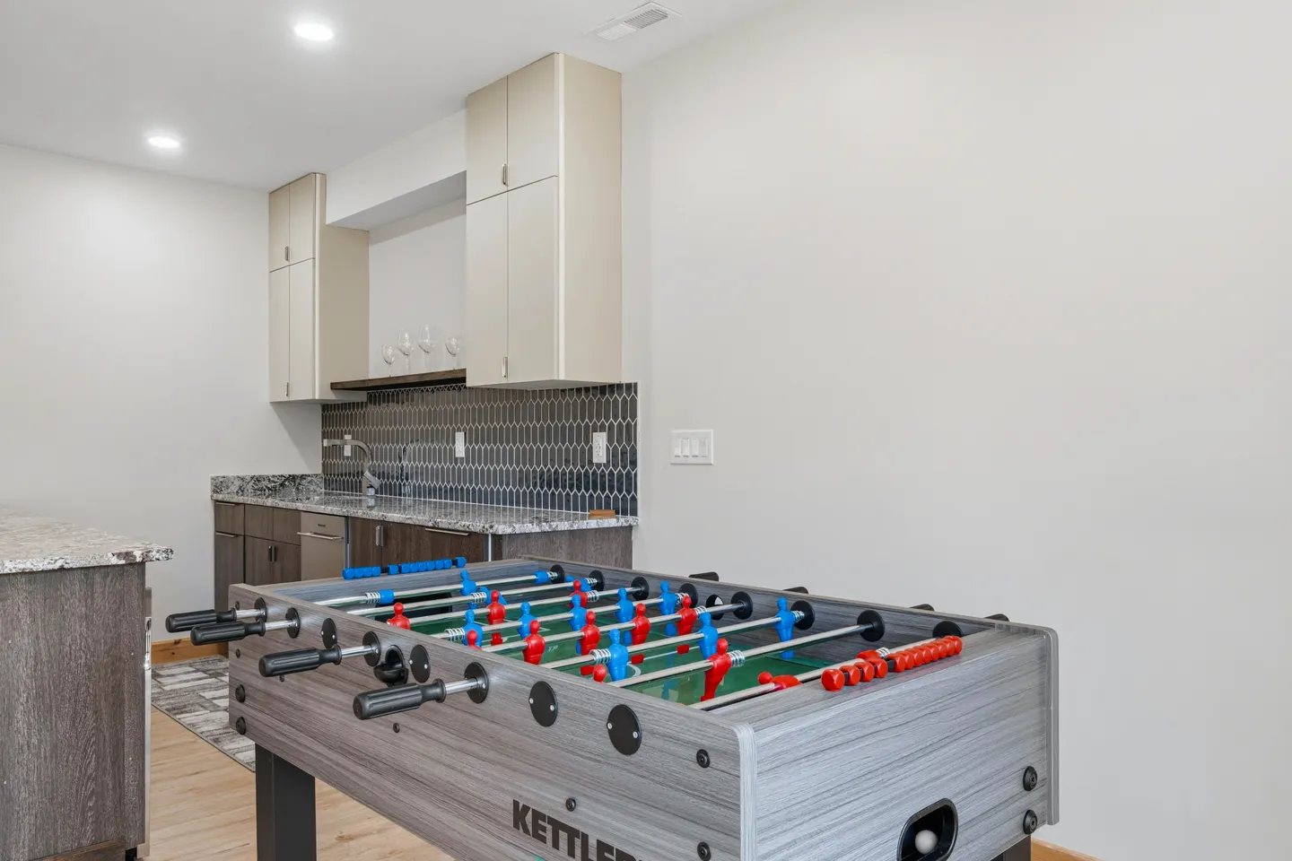 A foosball table positioned in a room with a wet bar featuring dark tile backsplash and stone counters in the background.