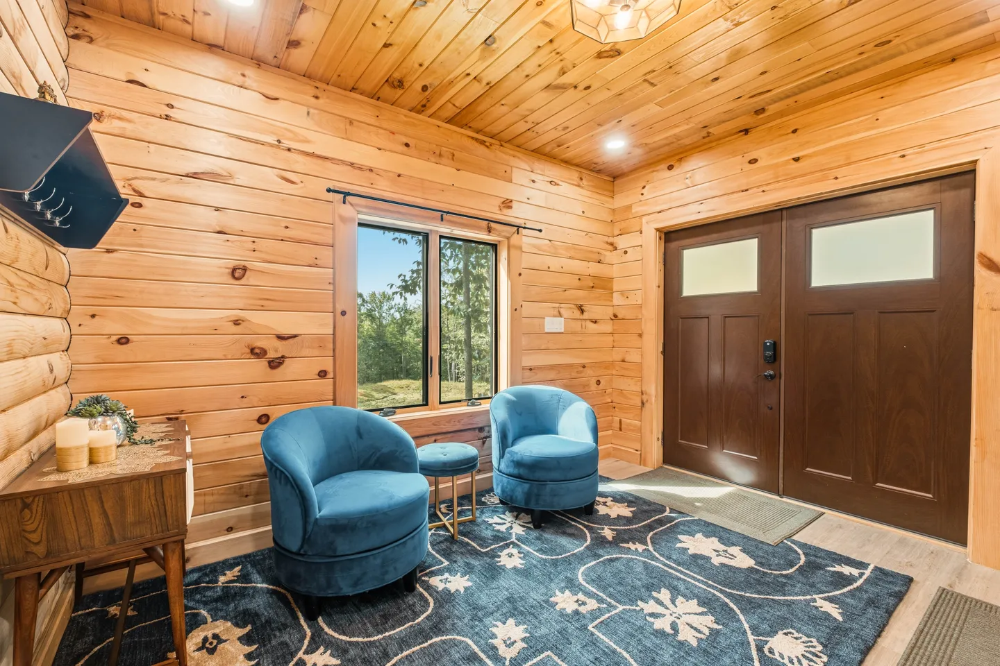 Foyer of a log cabin with wood-paneled walls, double front doors, a blue patterned area rug, and two blue velvet swivel armchairs.