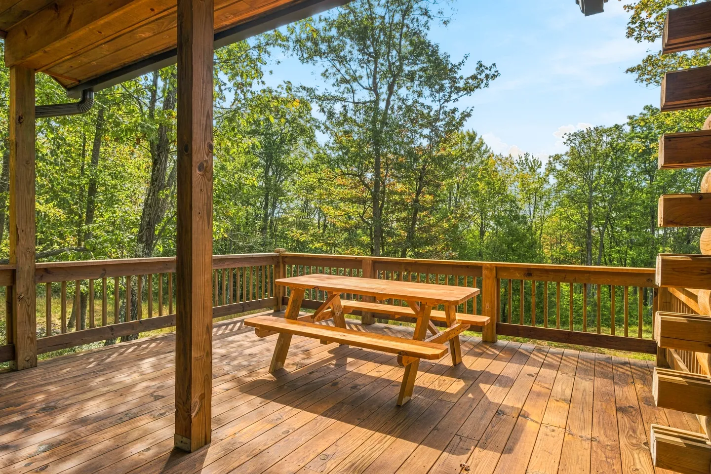 A wooden outdoor deck featuring a picnic table and railing overlooking dense green trees under a blue sky.