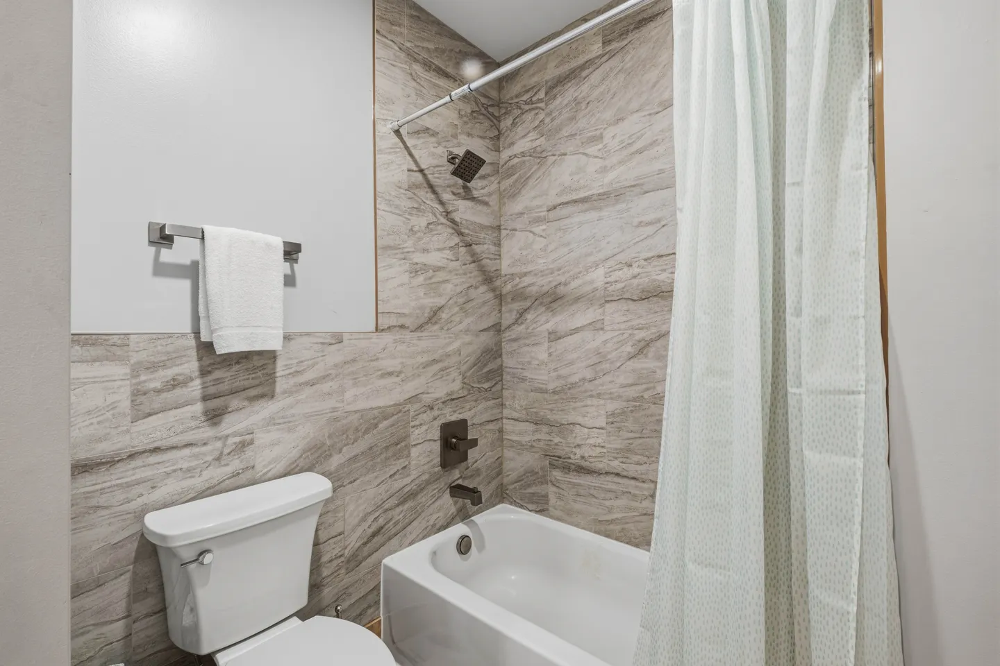 Bathroom showing a white toilet, white bathtub, dark fixtures, and patterned beige tile surround.