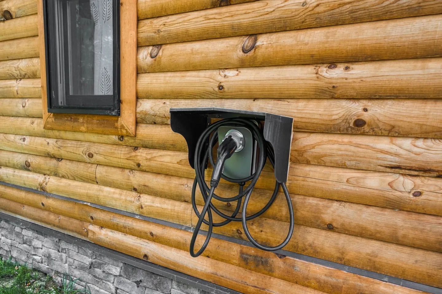 Electric vehicle charging station mounted under a protective hood on an exterior log cabin wall.