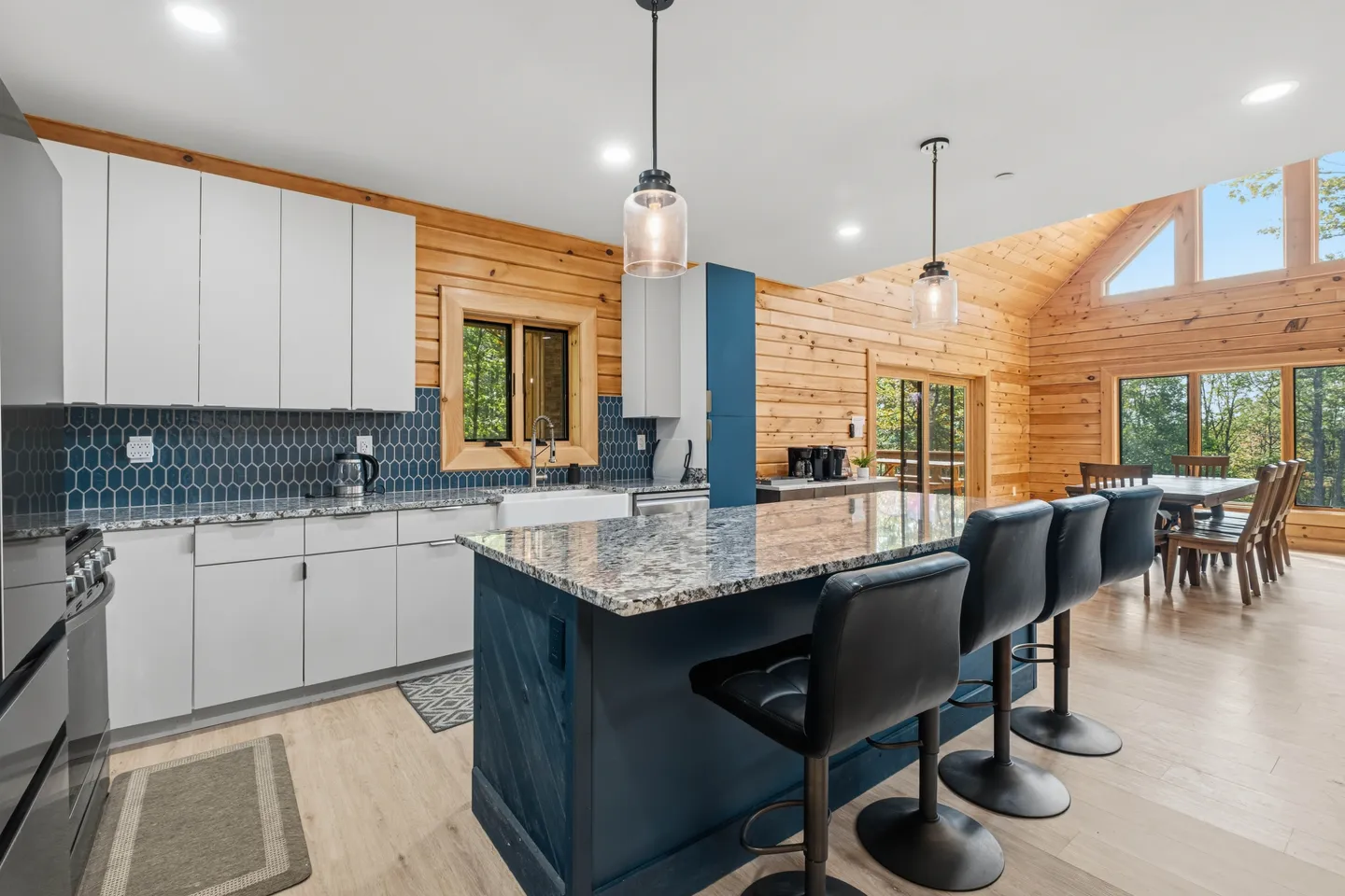 Kitchen with blue and white cabinets, a large island with black barstools, and wood-paneled log cabin walls leading to a dining area.